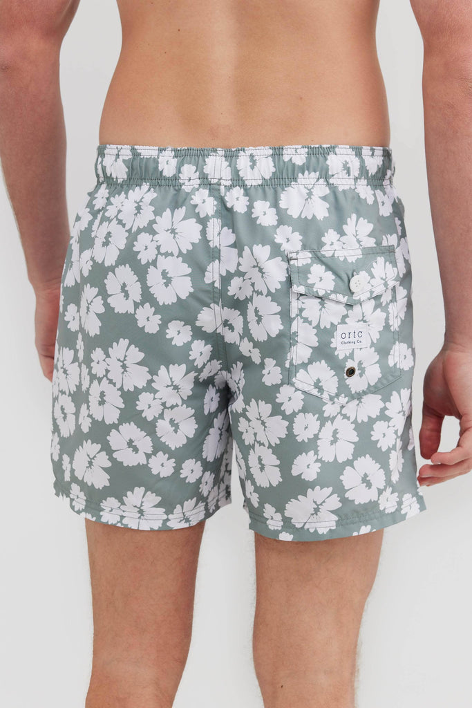 Back of model wearing sage board shorts with flowers on them. They have a pocket with ortc logo on them