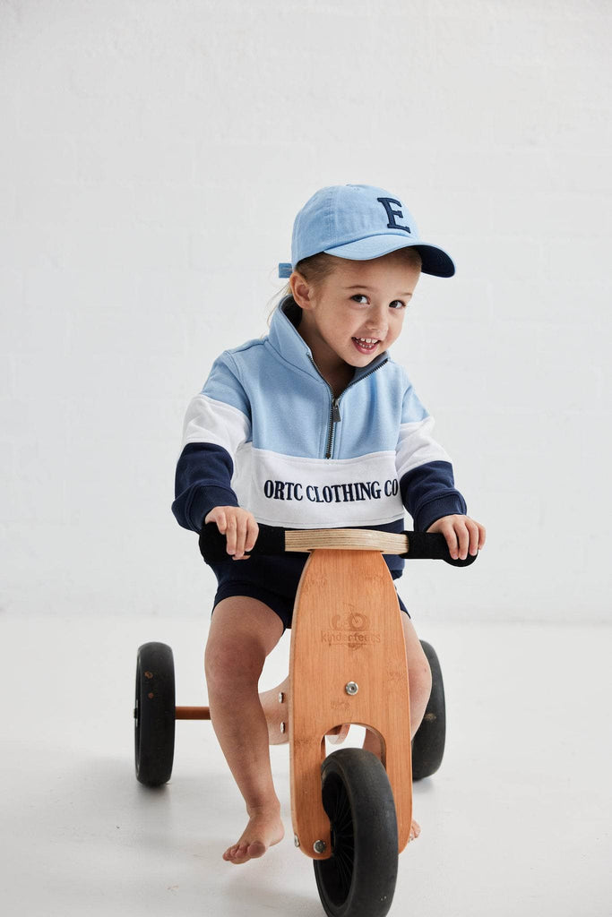 Child on a bike wearing quarter zip jumper that has a light blue top panel, white middle panel with navy embroidered ortc Clothing Co and navy base panel and a blue baseball cap with a navy E embroidered on it